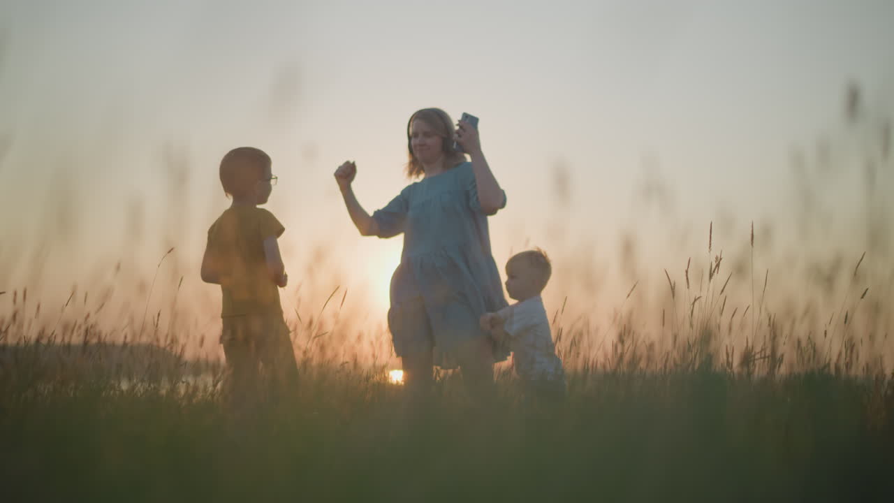 헤드을 착용한 파란색 드레스를 입은 여성이 스마트폰을 들고 해가 지는 동안 빛으로 빛나는 에서 두 아들과 함께 즐겁게 춤을 추고 있습니다.