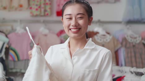 아시안 어머니가 아이들 중 한 명을 선택하고 에 옷을 걸고 미소 짓고 엄지손가락을 보여주고 부티크에서 쇼핑하는 동안 카메라에 제스처를합니다.