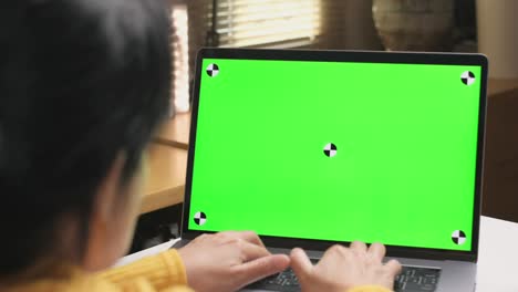 vista trasera de una mujer trabajando desde casa, escribiendo en una computadora portátil con pantalla verde y sentada en un escritorio blanco, trabajando en el concepto de hogar.