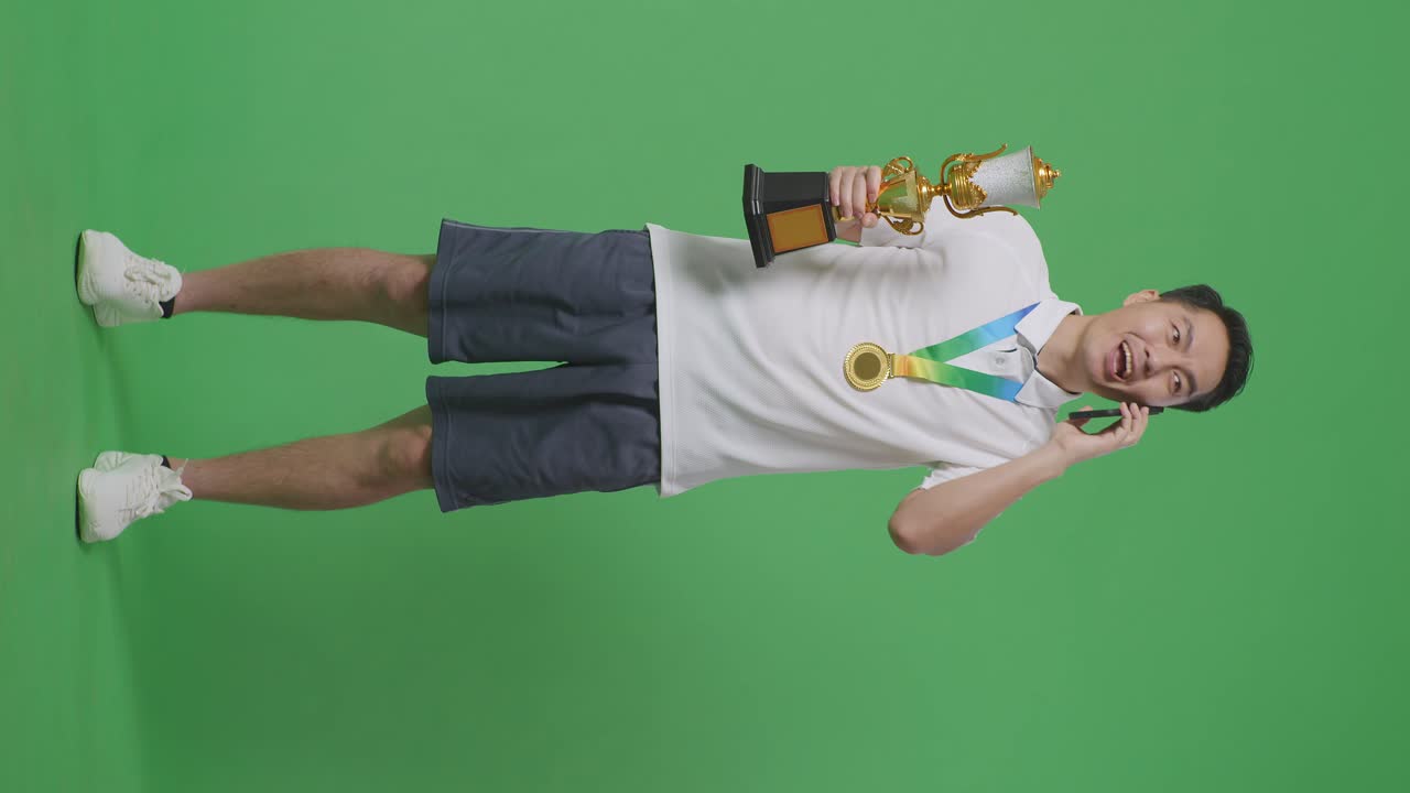 골드 트로피를 들고 스튜디오의 녹색 화면 배경에서 스마트폰으로 이야기하는 금메달을 가진 아시아 남자의 몸이 가득 차 있습니다.