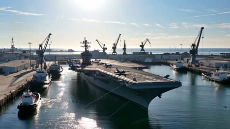 large aircraft carrier docked in a busy port with tugboats and fighter jets
