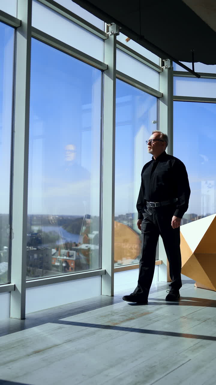 Adult man wearing black shirt and trousers walks along the panoramic window in the sunny office. Low angle view on the businessman looking at cityscape. Vertical video.