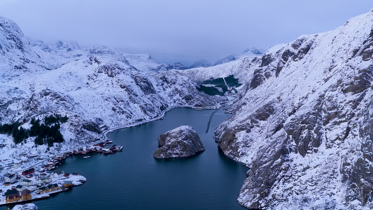 Filmische Luftaufnahme eines kleinen Fischerdorfes, eingebettet in einem verschneiten Fjord auf den Lofoten, Norwegen. Wunderschöne Winterlandschaft mit dunkelblauem Wasser und weißen Berggipfeln