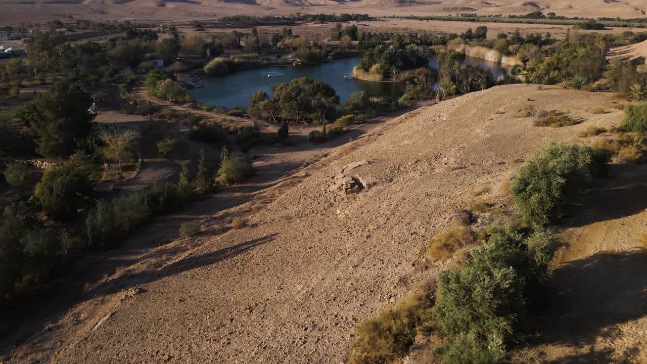 Drone push-in over Neot Smadar oasis, lush green trees, tranquil lake and dry desert hills under warm golden light.