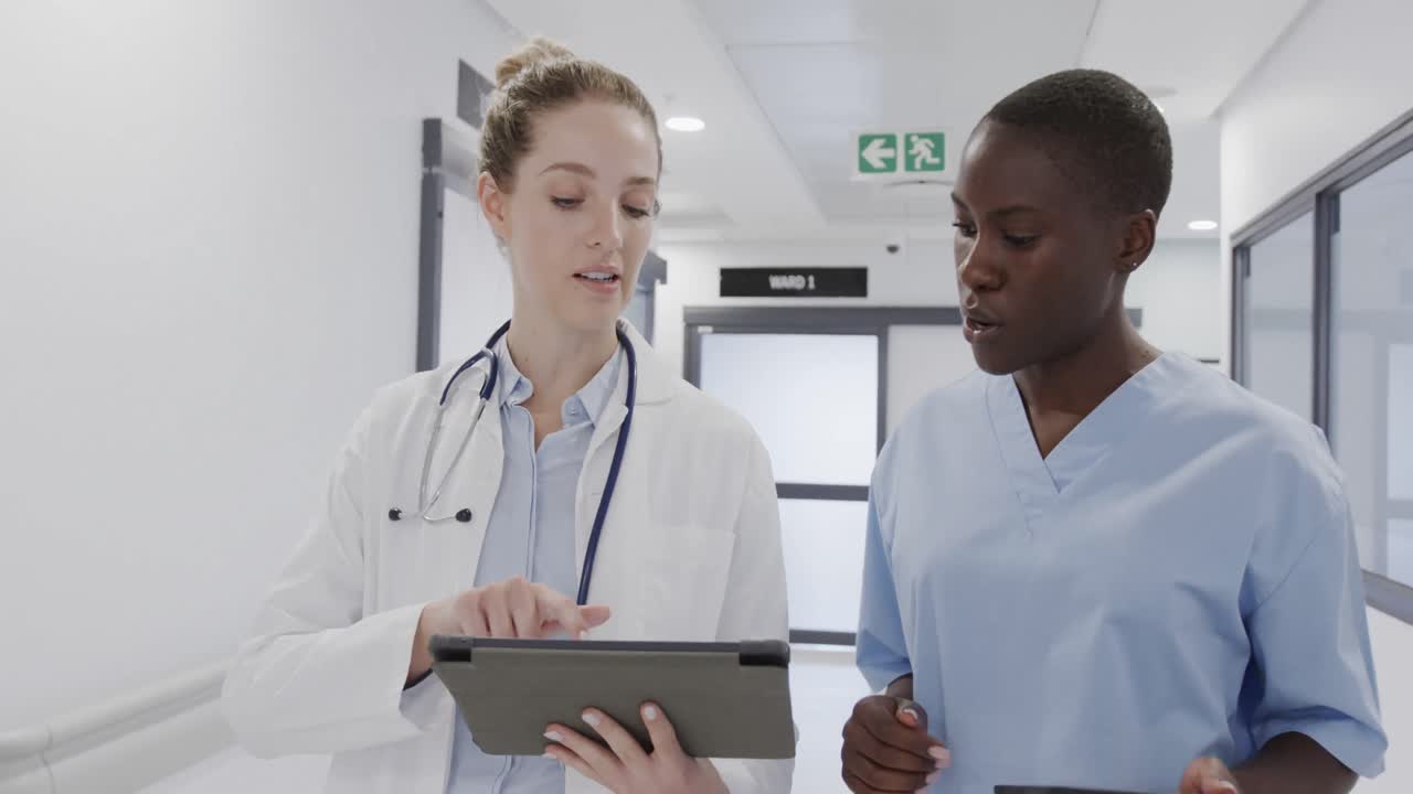 Diverse female doctor and nurse using tablet and discussing in hospital corridor, in slow motion