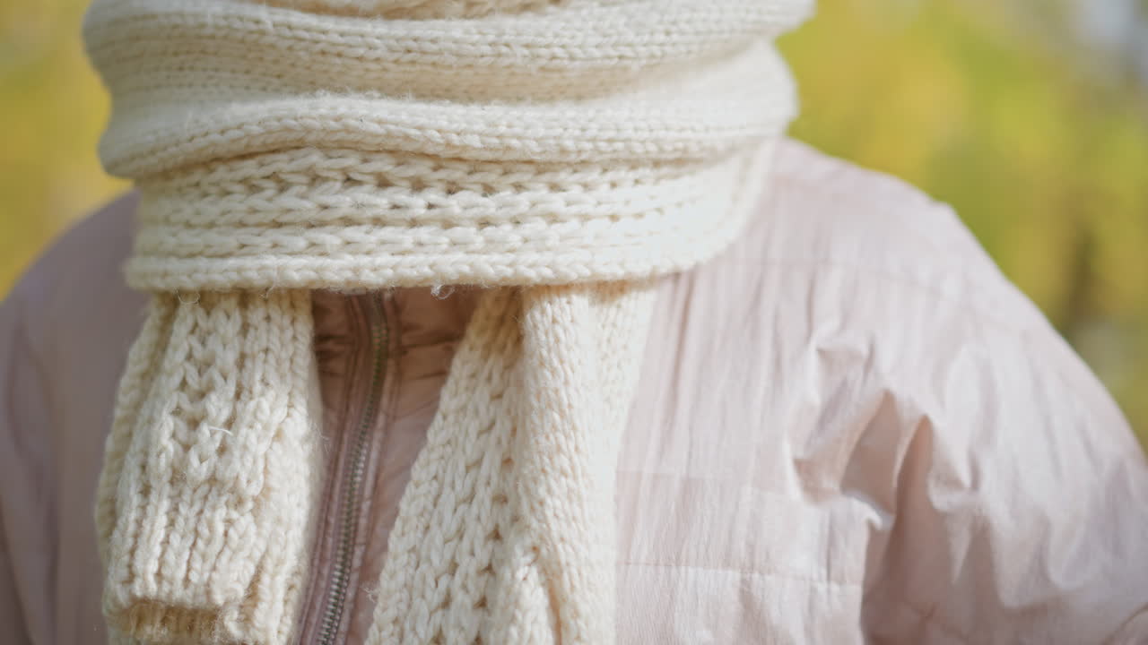 close up of woman adjusting chunky knit scarf around neck in autumn setting with blurred yellow foliage background showing delicate French manicure and textured wool accessory in cozy seasonal attire