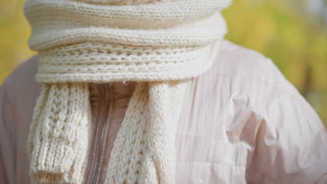 close up of woman adjusting chunky knit scarf around neck in autumn setting with blurred yellow foliage background showing delicate french manicure and textured wool accessory in cozy seasonal attire