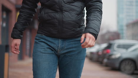 front lower view of vehicle owner wearing blue jeans and black winter coat walking along pavement with parked cars and urban buildings in background during cold city day while pedestrians stroll past