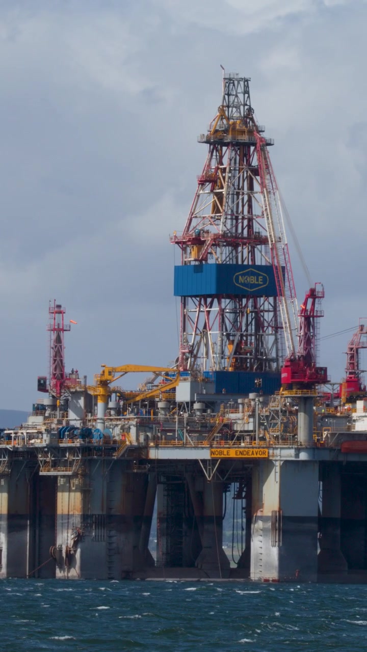 Stor offshore olieboreplatform i oprørt hav, overskyet dagslys, langsom sideværts kamerapanorering, industrielt miljø.