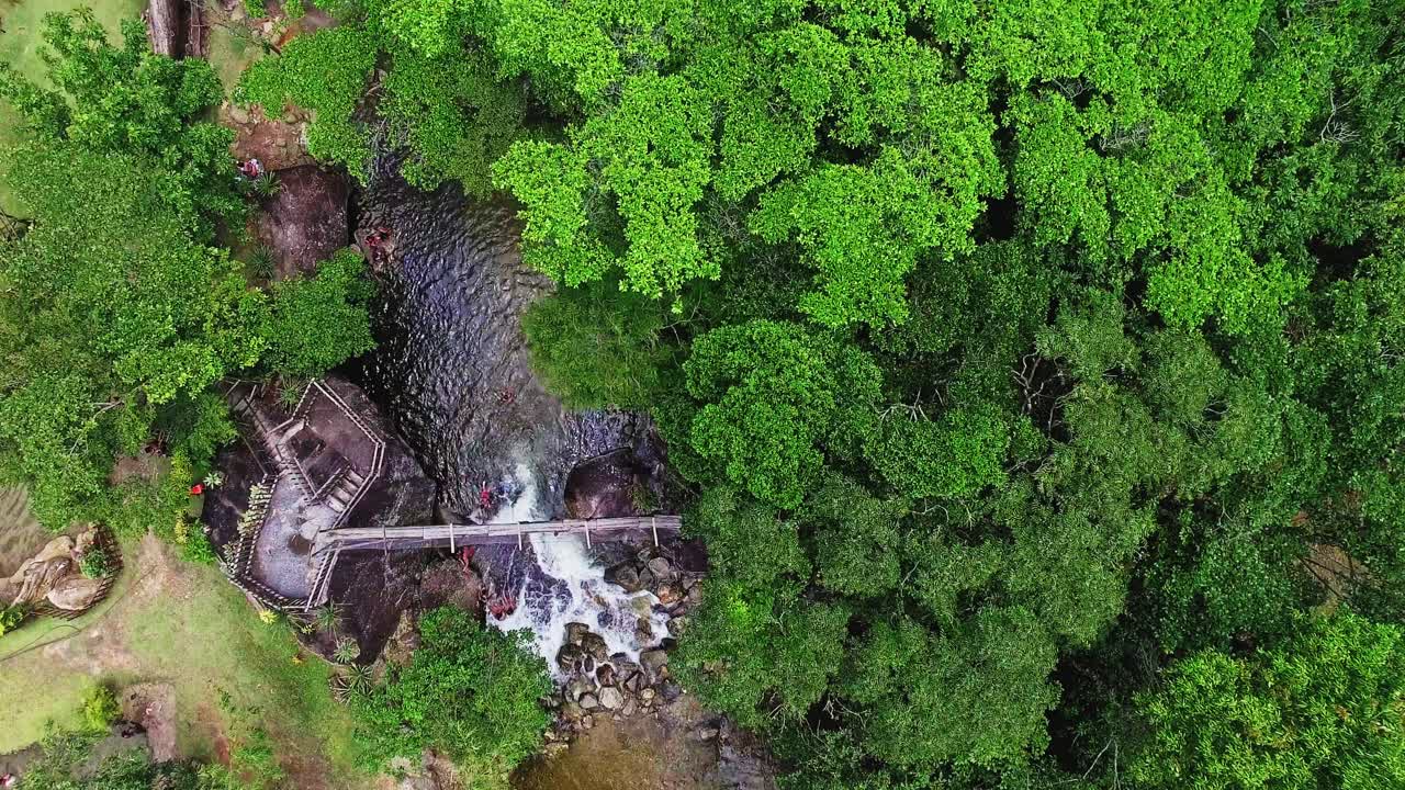 Beautiful 4K aerial view of a serene waterfall and river, surrounded by lush greenery. People swim and play in the water, with small wooden bridges crossing the river, perfect for nature