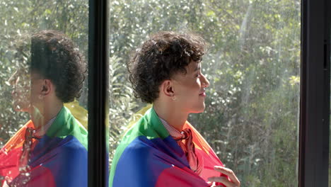 smiling young person wrapped in rainbow flag standing by window at home