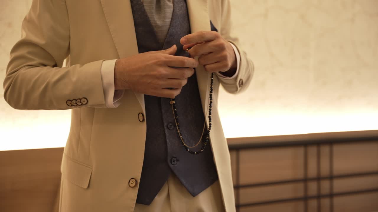 Close up of groom in beige suit holding black prayer beads with blue vest