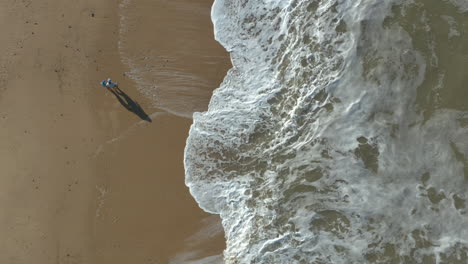 Aerial-shot-of-parent-and-child-social-distancing-while-walking-on-a-beach-with-waves-crashing