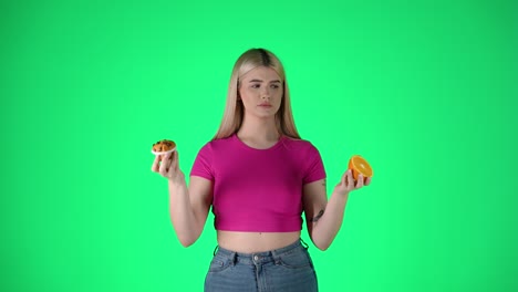 joven rubia con pastel y rebanada de naranja, elección entre comida saludable y no saludable, fondo verde tiro de estudio