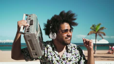 happy afro man having fun listening to music with wireless headphones and vintage boombox on the beach during summer time