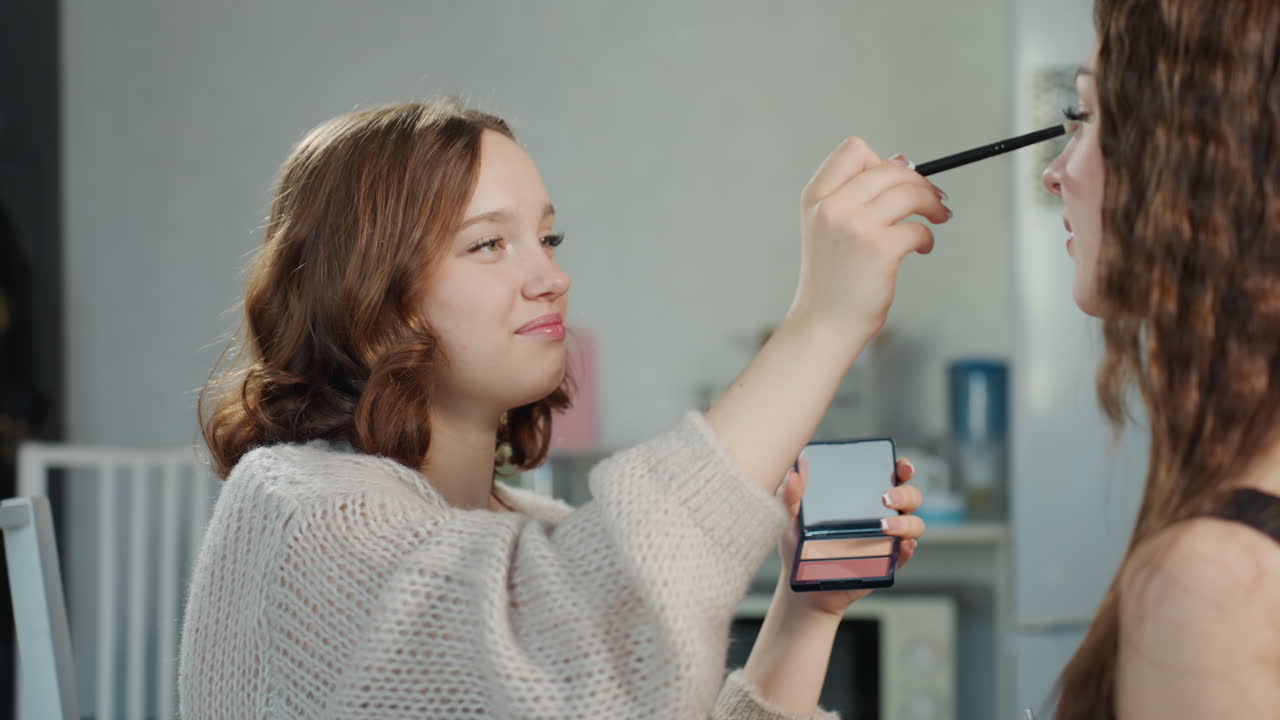 White Beauty Vlogger Testing Eyeshadow On Friend, Cozy Indoor Setup, Casual Recording Vibe, Palette And Brush In Hand, Smiling Interaction, Product Focus And Lighthearted Review Tone