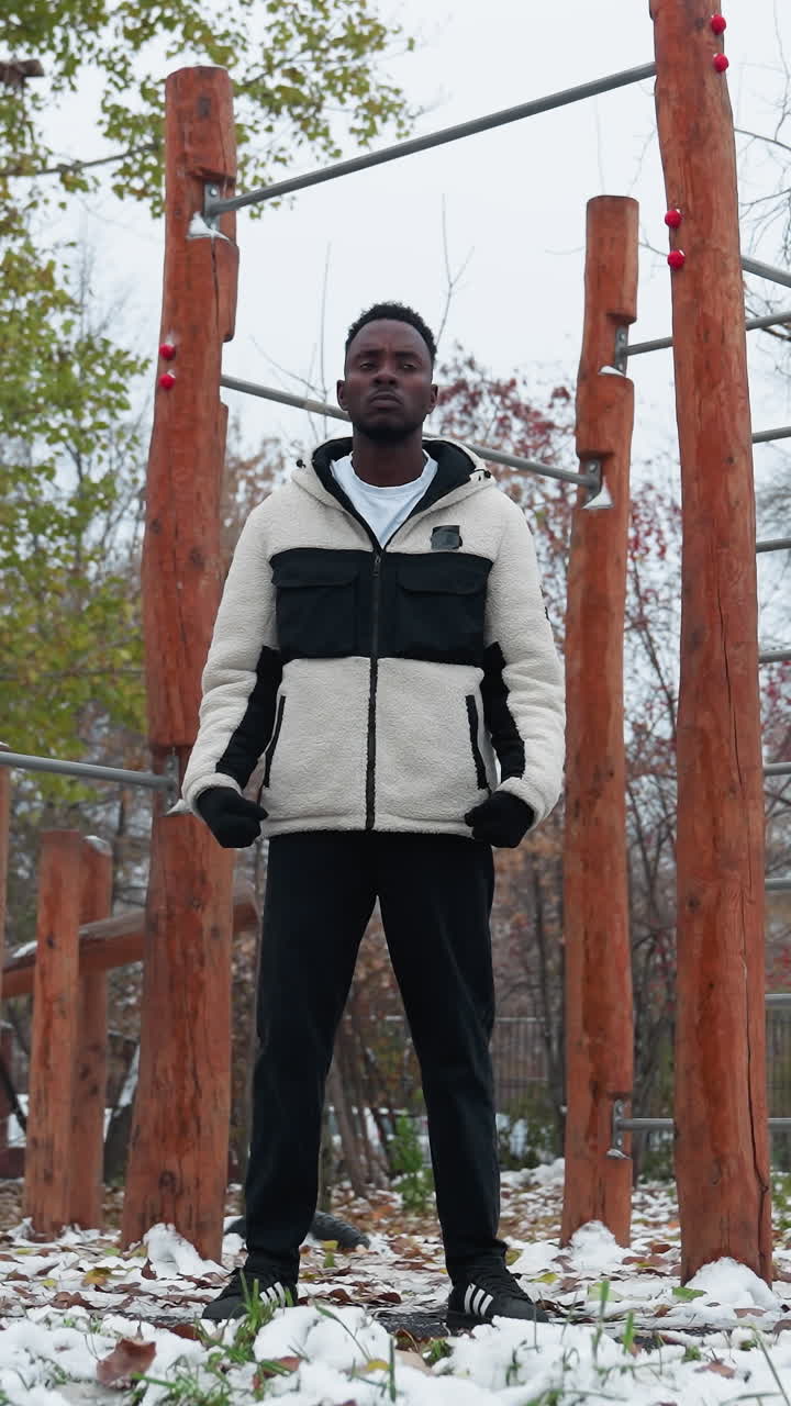 joven con guantes negros, chaqueta de invierno y pantalones negros realizando torsión de muñeca al aire libre, entorno de parque nevado con equipos de entrenamiento, hojas caídas y edificio residencial lejano