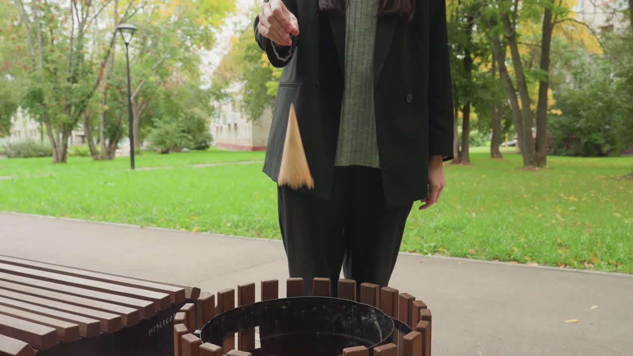Donna che si prepara a gettare le chiavi in un cestino vicino a una panchina e un sentiero, giacca casual, alberi autunnali e prato verde sullo sfondo, primo piano di una mano che lascia cadere un portachiavi sopra un cestino di legno, atmosfera pensierosa.