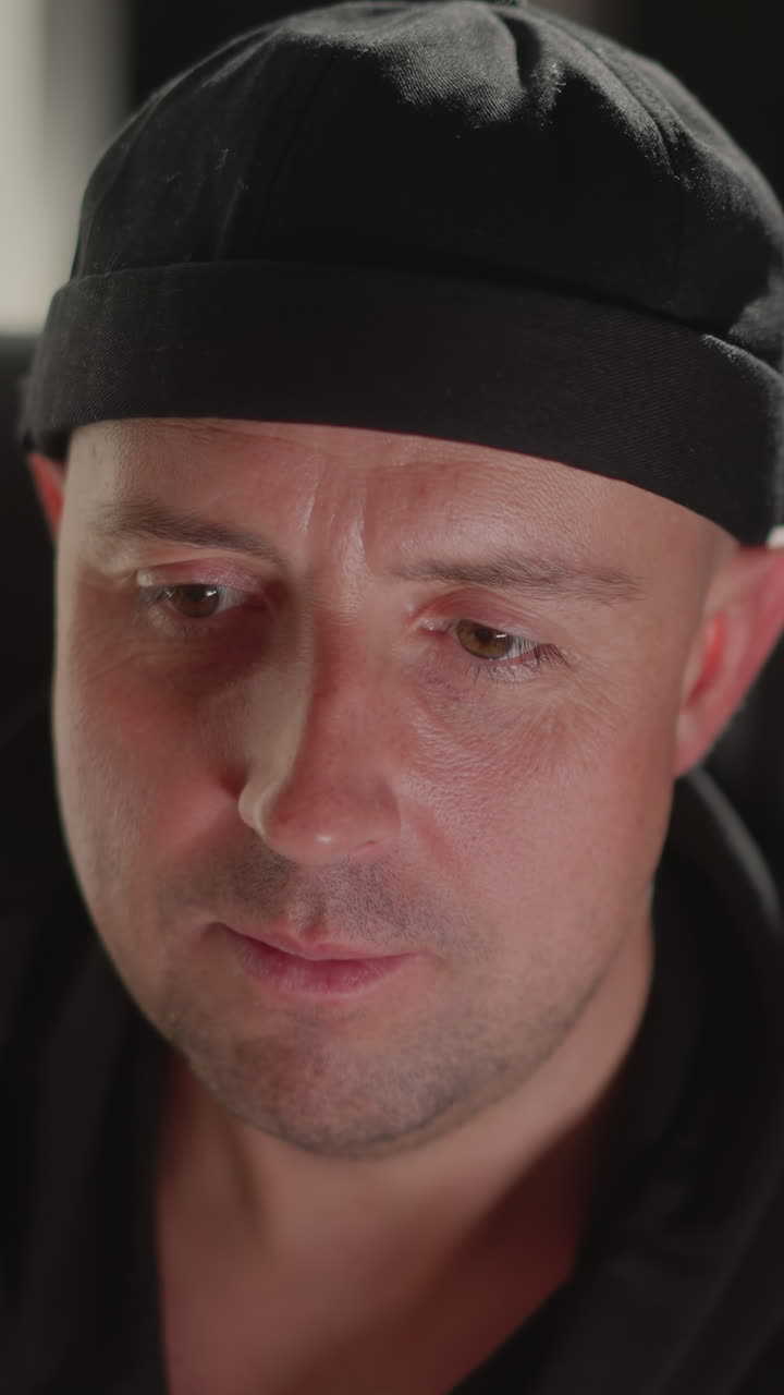 Close-up of a man wearing a black cap and black shirt, sitting on a black chair. His expression shows focus and concentration as he looks intently at something out of frame