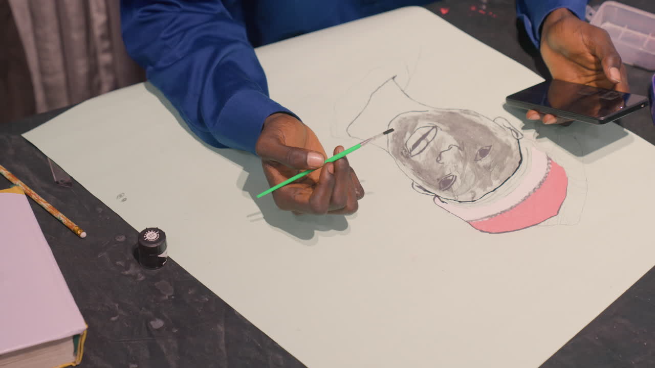 Closeup of black artist painting portrait, blue shirt, steady hands applying ink and brush strokes to grayscale face with pink cap, phone and palette nearby, calm studio atmosphere, focused sketching