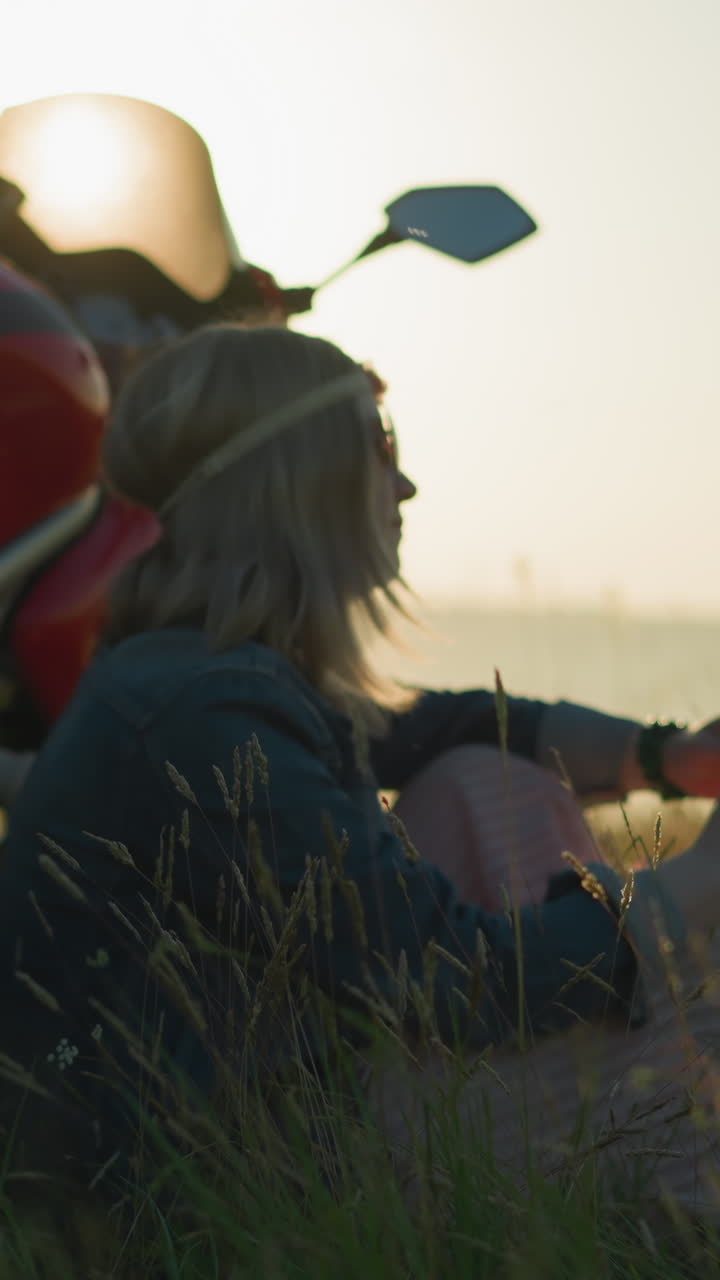 A woman with a flower headband and sunglasses sits in a grassy field beside a motorcycle, she thoughtfully plays with a strand of grass in her hand, lost in thought, as the sun sets behind her