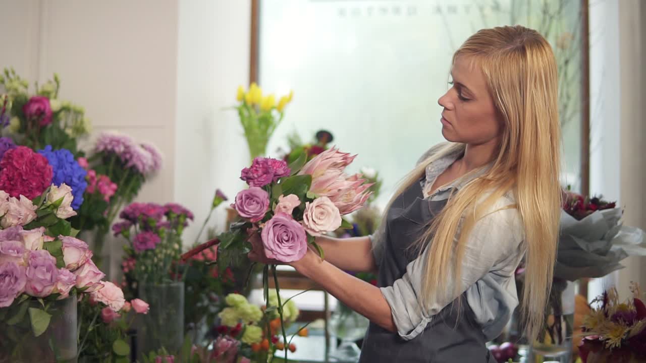 боковой вид молодой привлекательной женщины в фартуке, работающей в цветочном выстреле и устраивающей букет цветов. она выглядит очень