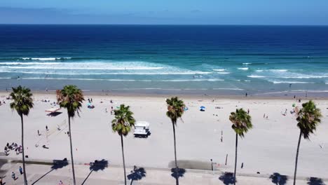 Aerial-of-a-drone-flying-along-palm-trees-with-the-blue-ocean-in-the-background-on-a-sunny-day-and-people-walking-on-the-beach