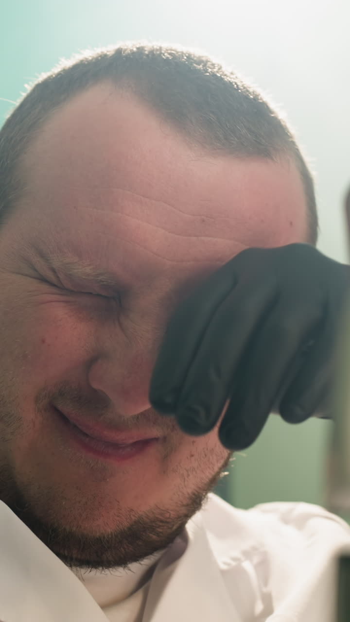 una vista de cerca de un científico con una bata blanca de laboratorio estremecéndose y usando sus manos para limpiar sus ojos, mientras examina muestras a través de un microscopio en un laboratorio