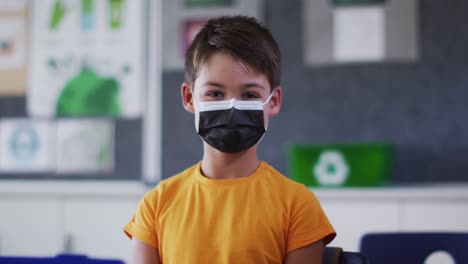 Portrait-of-mixed-race-schoolboy-wearing-face-mask,-sitting-in-classroom-looking-at-camera