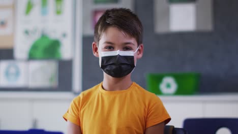 portrait of mixed race schoolboy wearing face mask, sitting in classroom looking at camera