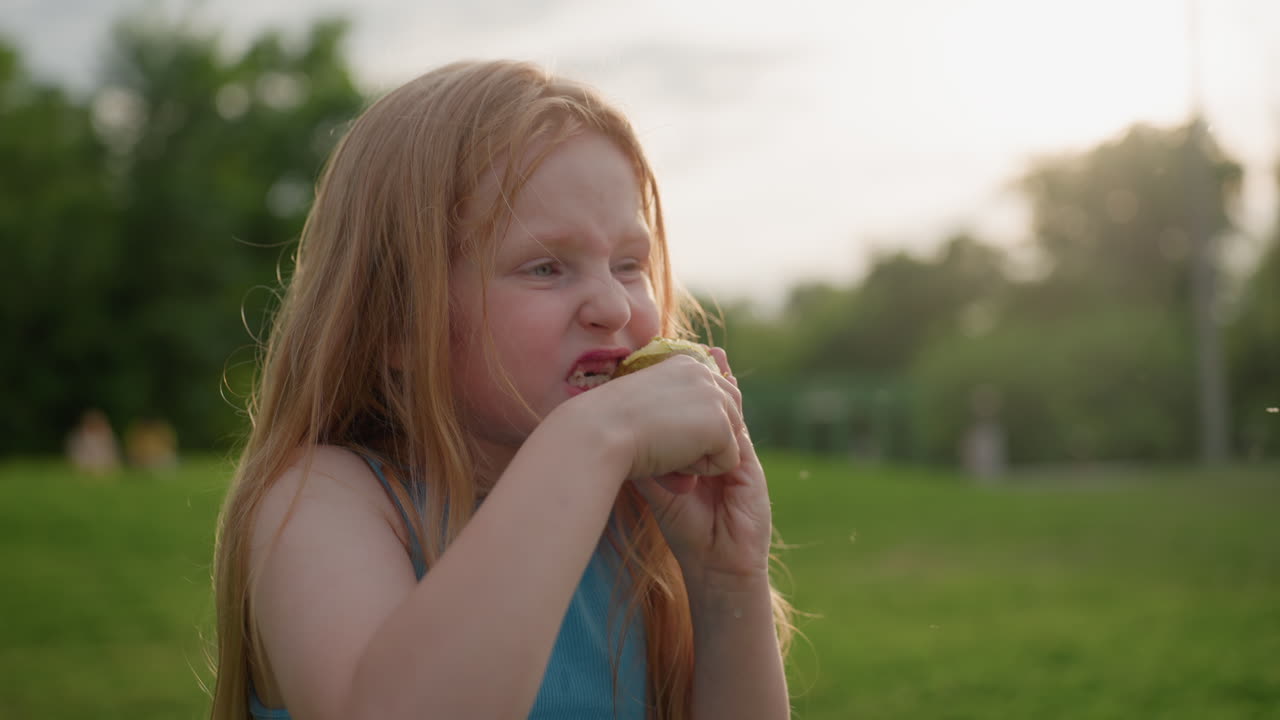young girl in park trying to cut pear with side teeth while chewing with determined expression, sunlight backlight, green grass background, playful struggle showing childhood curiosity