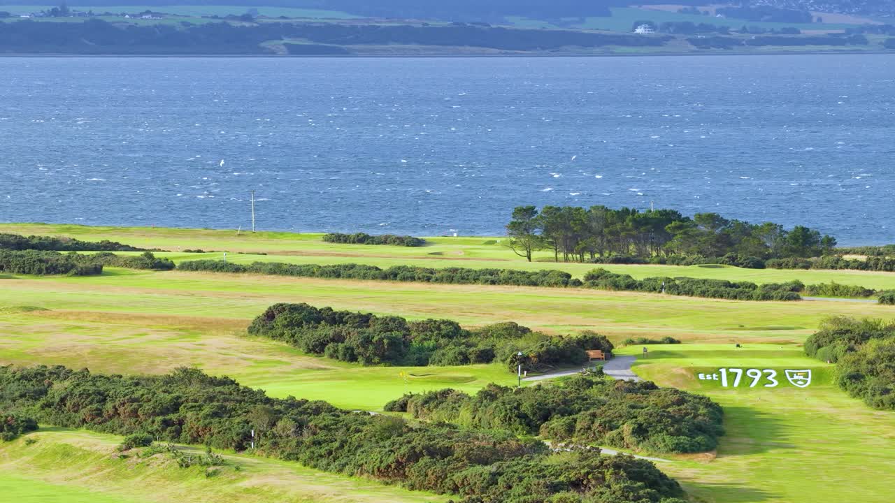 Wide shot pans over lush seaside golf course, vibrant greenery, blue water, natural daylight