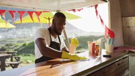 hombre afroamericano con delantal y guantes limpiando el camión de comida con spray desinfectante