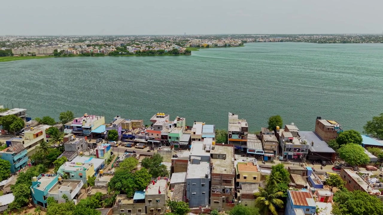 Vista aerea con drone di case colorate sul lungomare e densi edifici residenziali che si affacciano su un vasto lago, con una costa urbana distante e uno skyline sfocato a Chennai, India.