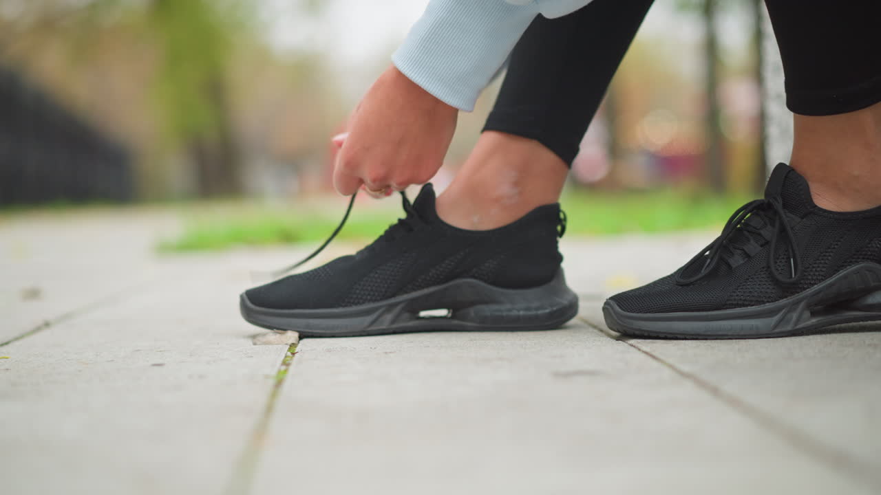 Close-up shot of lady tying right leg shoelace in black sneakers, preparing for outdoor workout or run, focus on hands tying shoes on sidewalk, active lifestyle, fitness preparation in park