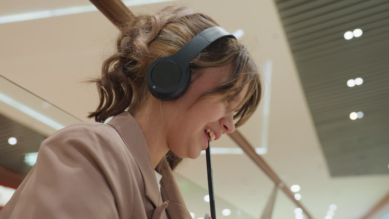 Mujer joven en una lluvia de ideas; mujer concentrada con auriculares grabando sus pensamientos con calma; mujer joven con auriculares en un centro comercial capturando ideas en una libreta reflexivamente durante un ensayo