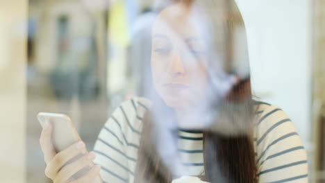 die kamera fokussiert eine brünette frau durch das fenster, die kaffee trinkt und auf dem smartphone sms schreibt, während sie an einem tisch in einem café sitzt