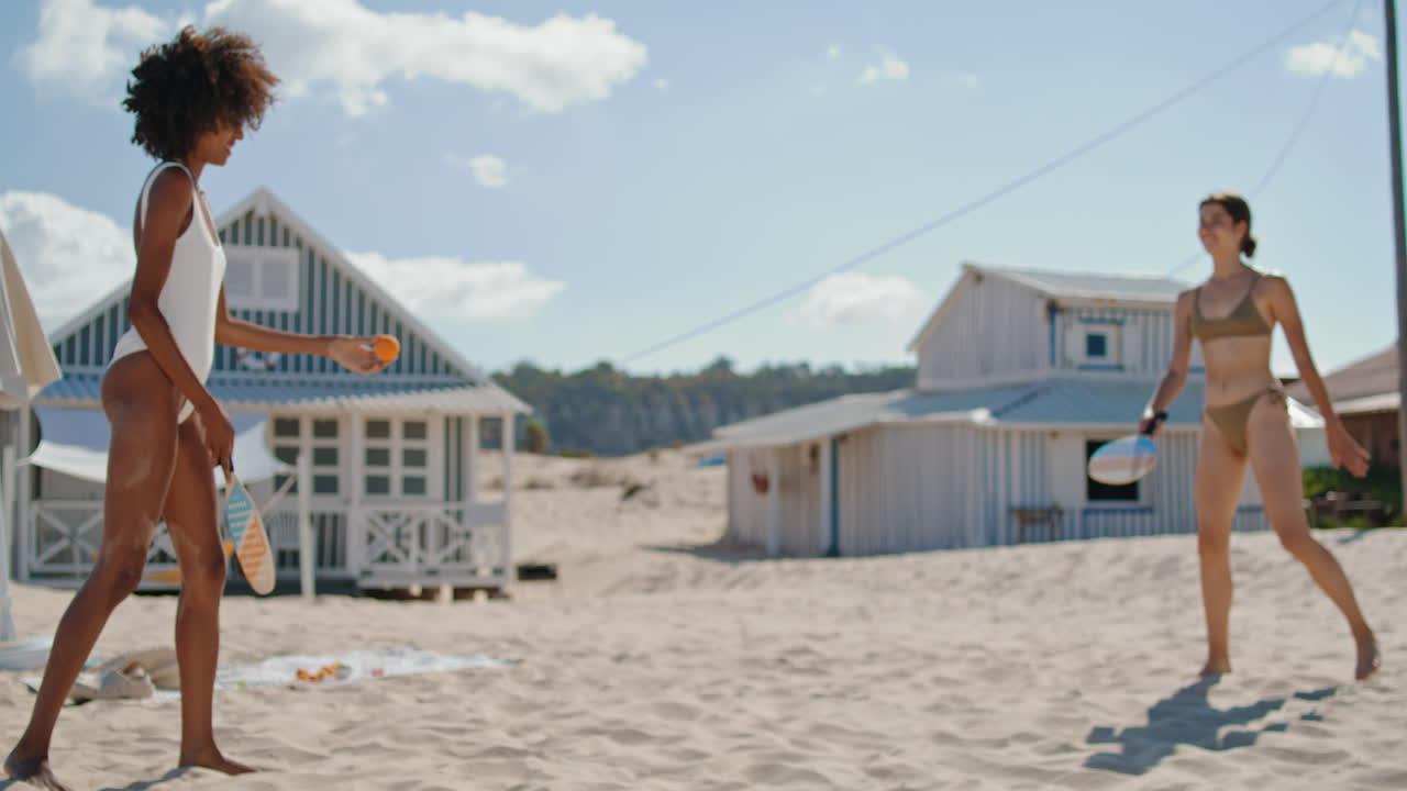 모래 해변에서 테니스를 하는 즐거운 파트너들. 비키니를 입고 재미있는 행복한 소녀들