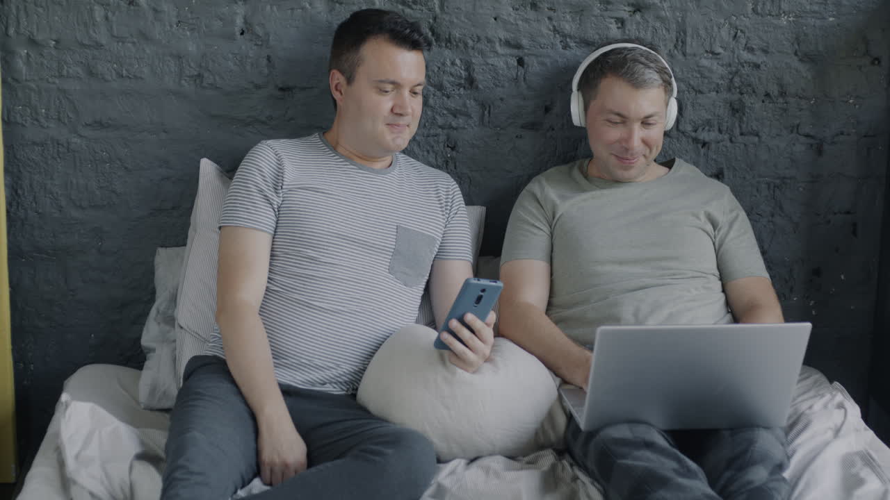 Two Men Relaxing on a Bed with a Laptop and Phone