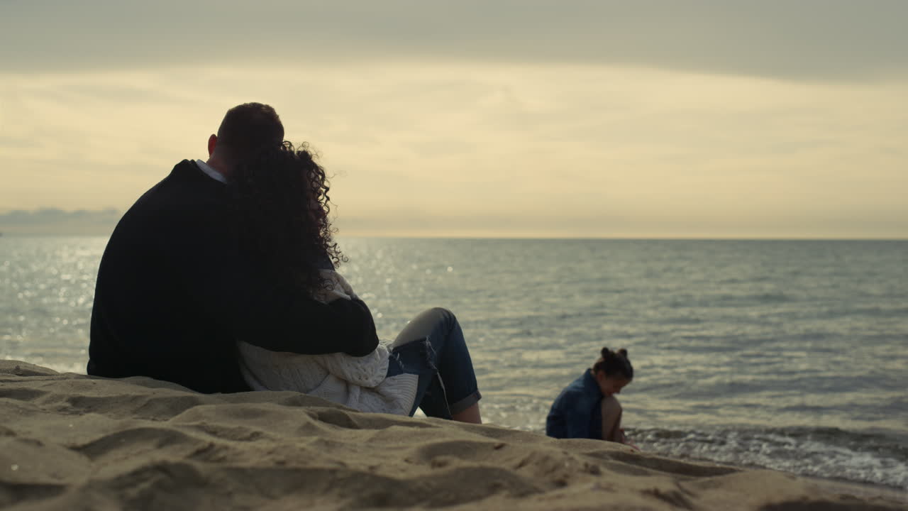 해가 지는 해변을 즐기는 편안한 사람들이 모래 해안에 앉아 가족들이 냉정하게 지내고 있습니다.