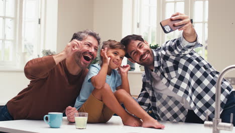 same sex family with two dads pulling faces for selfie in kitchen with son sitting on counter