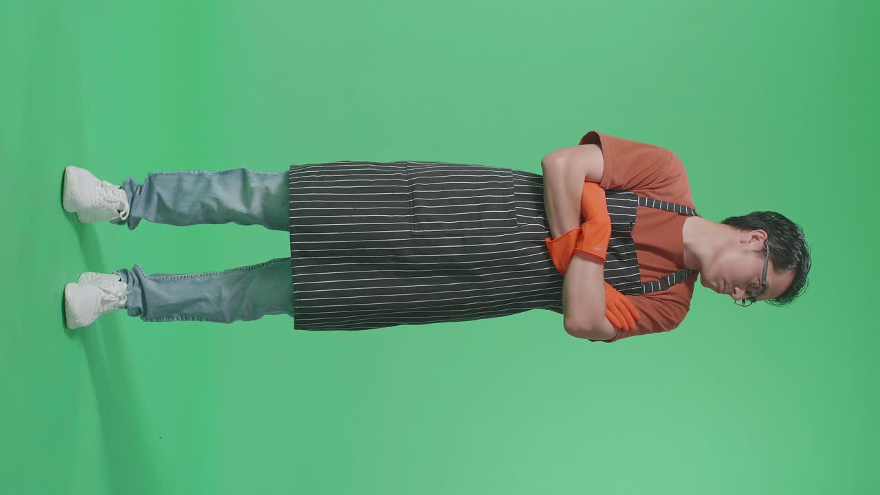 화가 난 아시아 남성 집주인이 팔을 고 녹색 스크린 스튜디오에 서서 주위를 둘러보고 있습니다.