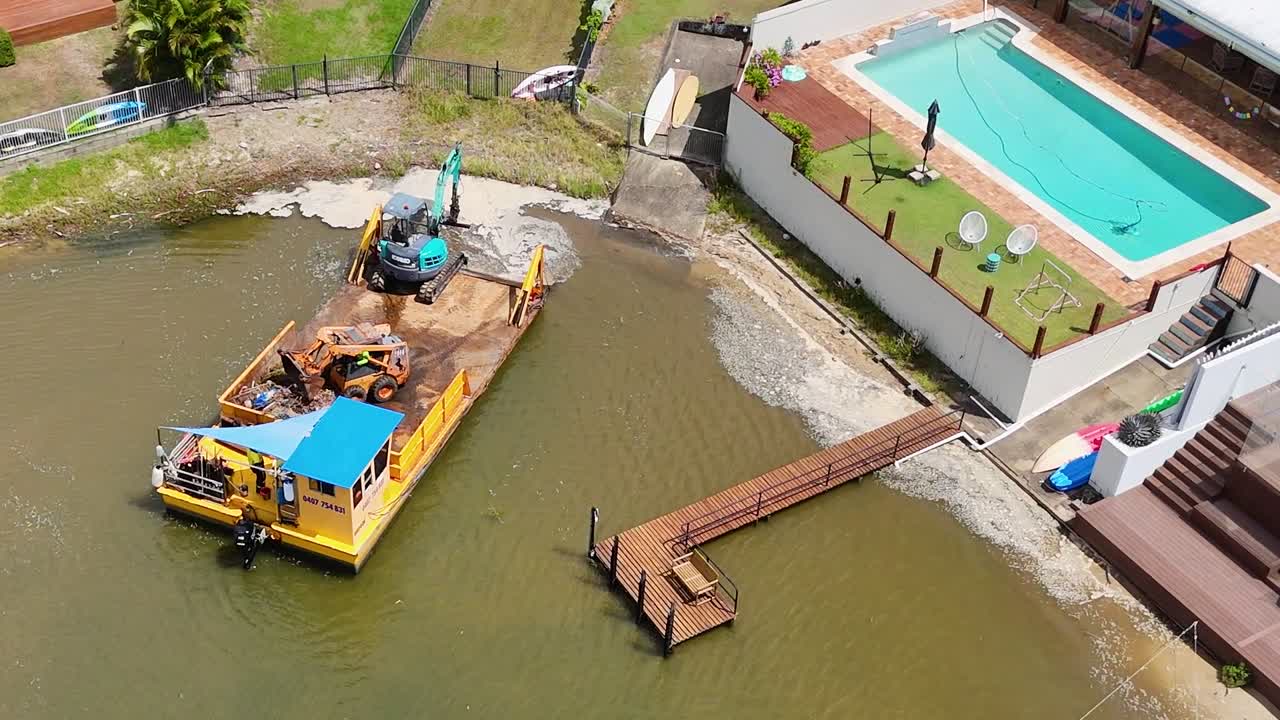 imágenes de drones capturan una barcaza y una grúa trabajando en un canal cerca de casas de lujo con piscinas