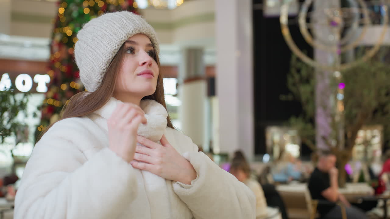 Elegant woman wearing a fluffy winter coat and putting on a knitted head warmer in a cozy indoor setting, blurred background features seated people and festive decor with warm bokeh lighting
