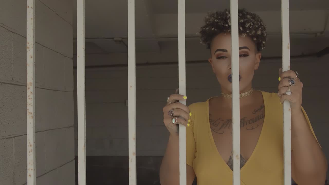 Latin woman with visible tattoos on her chest approaches prison bars. Zoom out