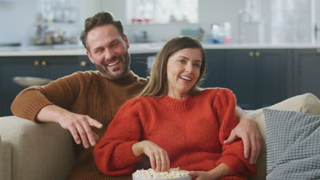 couple sitting on sofa with popcorn laughing watching comedy on tv together