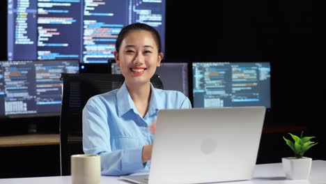 asian female programmer smiling to camera and cross arm while writing code by a laptop using multiple monitors showing database on terminal window desktops in the office