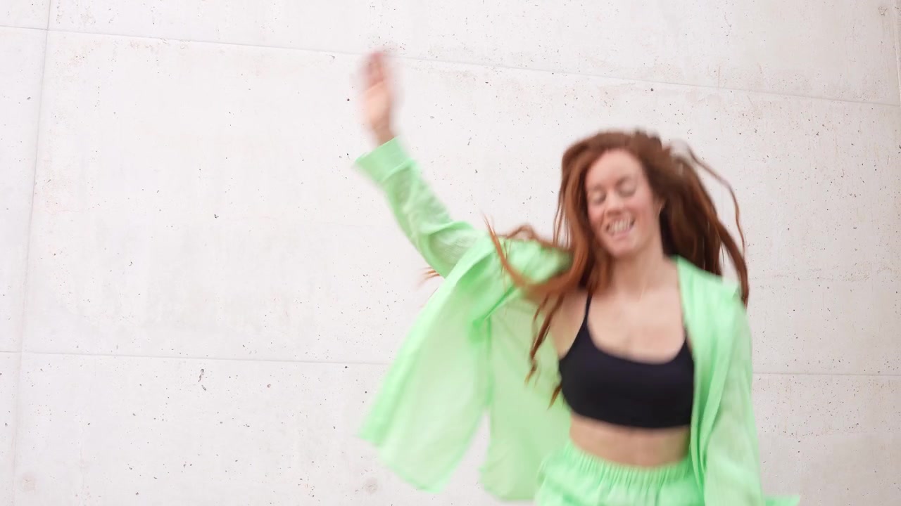A young woman clad in green dancing happily next to a concrete wall in an urban setting