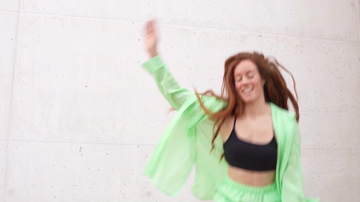 A young woman clad in green dancing happily next to a concrete wall in an urban setting