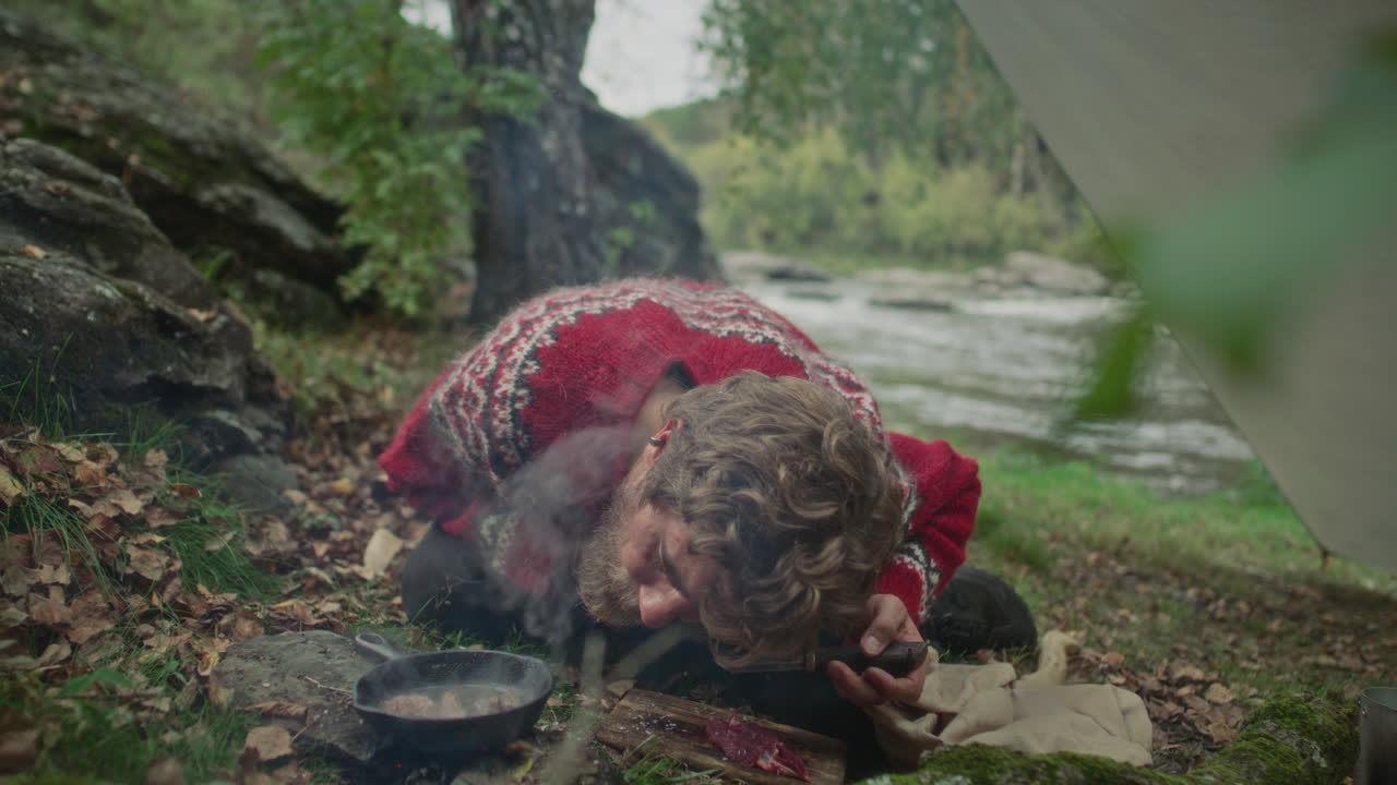 Man Cooking Meat over Campfire beside River in Forest